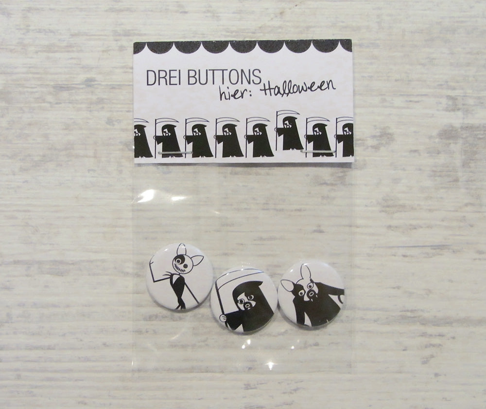 Handgefertigte Halloween-Buttons mit gruseligen Figuren in Schwarz-Weiß. Set aus drei Anstecknadeln mit verschiedenen Motiven: Sensenmann, Geist und Skelett. Verpackung zeigt Reihe stilisierter Sensenmänner. Perfekt für Halloween-Kostüme und gruselige Outfits.