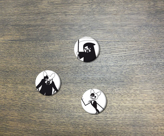 Drei runde Halloween-Buttons mit schwarzen Gruselgestalten auf weißem Hintergrund, liegend auf grauer Holzoberfläche. Die Buttons zeigen einen Sensenmann, eine Hexe und ein Skelett, perfekt für festliche Dekoration oder modische Akzente.