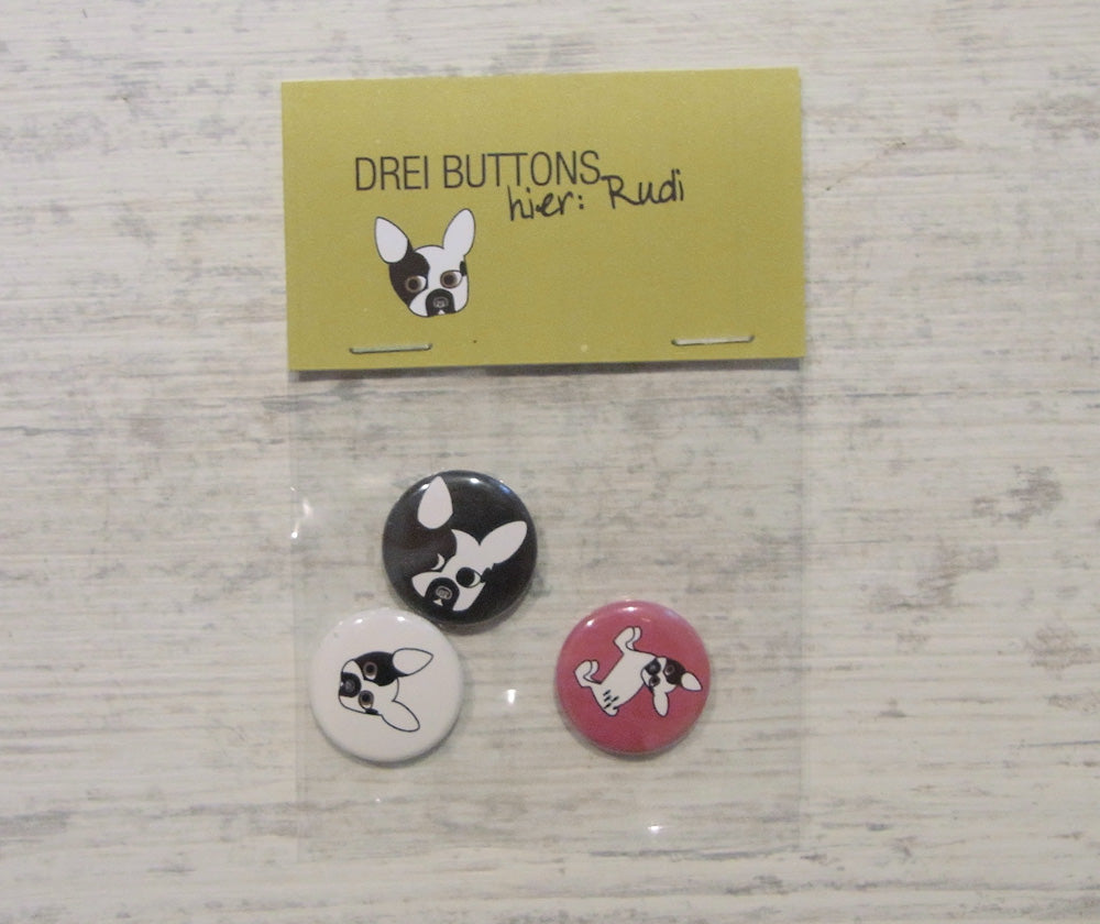 Handgefertigtes Button-Set mit drei Rudis: schwarz-weißer Hundekopf auf schwarzem und weißem Hintergrund, rosa Button mit geflecktem Hund. Verpackt in durchsichtiger Tüte mit gelbem Etikett "DREI BUTTONS hier: Rudi".