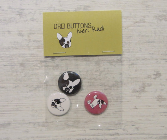 Handgefertigtes Button-Set mit drei Rudis: schwarz-weißer Hundekopf auf schwarzem und weißem Hintergrund, rosa Button mit geflecktem Hund. Verpackt in durchsichtiger Tüte mit gelbem Etikett "DREI BUTTONS hier: Rudi".