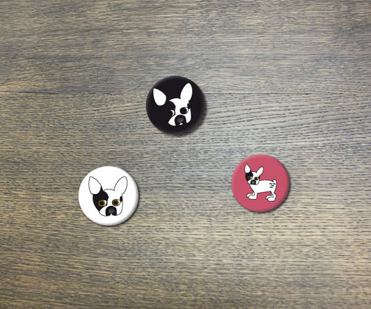 Handgefertigtes Button-Set mit drei Rudi-Motiven: schwarzer Hintergrund mit weißem Kopf, weißer Hintergrund mit schwarzem Kopf und pinkfarbener Hintergrund mit geflecktem Hund. Anstecknadeln auf Holzuntergrund präsentiert.