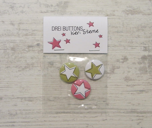 Handgefertigtes Dreier-Set von Buttons mit Sternen-Motiv in grün, weiß und pink. Verpackt in einer durchsichtigen Tüte mit einer Karte, auf der "DREI BUTTONS wer sterne" und kleine rosa Sterne abgebildet sind. Präsentiert auf einem hellen Holzuntergrund.