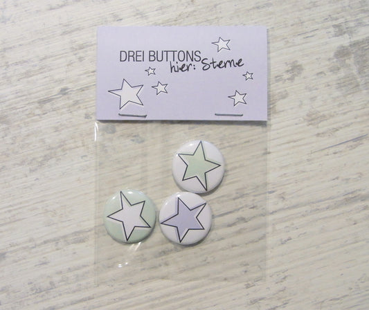 Handgefertigtes Dreier-Set von Buttons mit Sternenmotiv in weiß, grün und lila. Verpackt in einem durchsichtigen Beutel mit einer lila Karte, auf der "DREI BUTTONS hier: Sterne" und kleine Sternzeichnungen zu sehen sind.