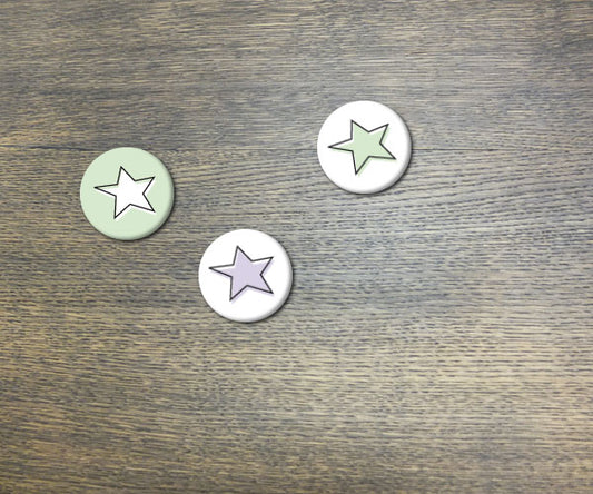 Drei handgefertigte Buttons mit Sternen-Motiv auf Holzuntergrund. Ein hellgrüner Button mit weißem Stern, ein weißer Button mit grünem Stern und ein lila Button mit weißem Stern. Jeder Button ist einzigartig gestaltet und Teil des DREI BUTTONS Sterne Sets.