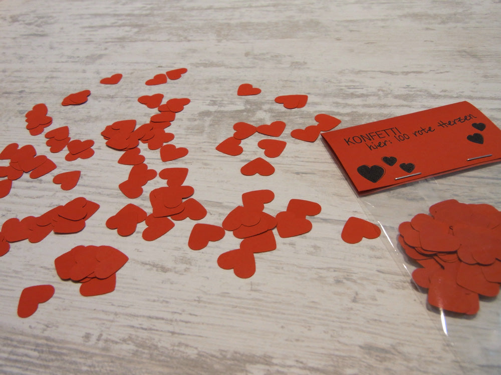 Handgestanztes rotes Herzkonfetti aus Papier für Hochzeit oder Valentinstag, verstreut auf hellem Holzuntergrund. Produktkarte mit KONFETTI-Aufschrift und Herzdesign sichtbar.