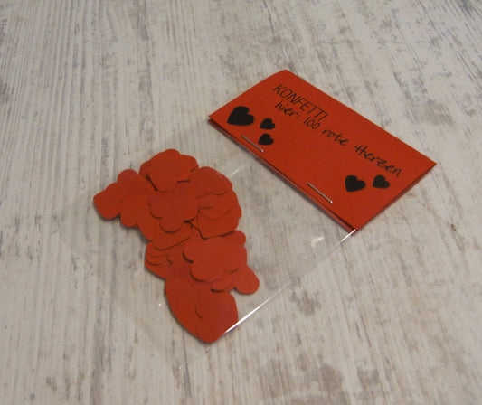 Handgestanztes rotes Herzkonfetti aus Papier neben der Produktverpackung auf hellem Holzuntergrund. Liebevolle Dekoration für Hochzeit oder Valentinstag, individuell gefertigt von Frau Hamster.